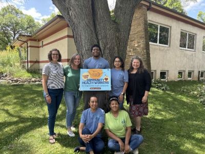 Seven person group standing in front of tree on a sunny day.