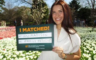 Medical student Alyssa Coley poses outside on a sunny day in front of a patch of tulips with a sign that shows her matching to DFMCH.