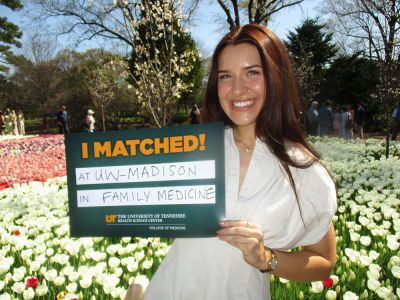 Medical student Alyssa Coley poses outside on a sunny day in front of a patch of tulips with a sign that shows her matching to DFMCH.